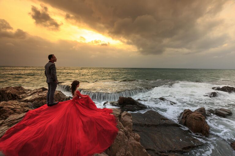 woman in red gown and man in gray suit standing and sitting on boulders in front of ocean