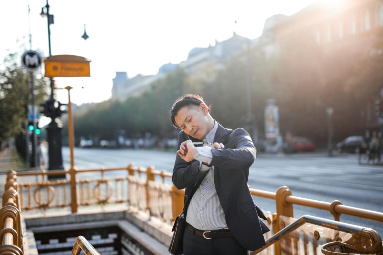 photo of man in blue blazer and striped shirt standing by stairs while on the phone and looking at his watch